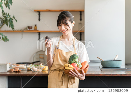 Young Asian woman in apron holding vegetables, avocado in kitchen 126908099