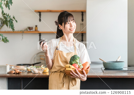 穿著圍裙的年輕亞洲女性在廚房裡拿著蔬菜和酪梨 126908115