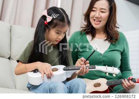 Parent-Child Bonding Through Music. A mother teaches her daughter to play the ukulele at home. 126908172