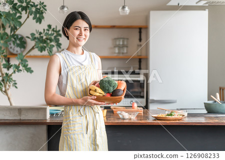 一位年輕的亞洲女性/家庭主婦，留著短髮，繫著圍裙，在廚房裡拿著蔬菜 126908233