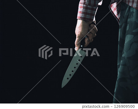 Male hand in plaid shirt and camouflage pants holding large kitchen knife downward in dramatic low light on dark background Male hand in plaid shirt and camouflage pants holding large kitchen knife downward in dramatic low light on dark background 126909500