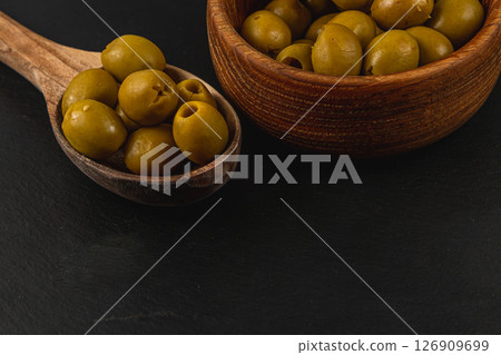 Green olives in a wooden spoon and bowl on a black surface 126909699