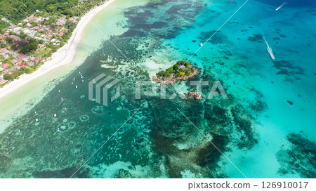 A small, lush island sits in shallow turquoise waters, surrounded by coral reefs and close to a white sandy shore. Praslin, Seychelles. A small, lush island sits in shallow turquoise waters, surrounded by coral reefs and close to a white sandy shore. Praslin, Seychelles. 126910017