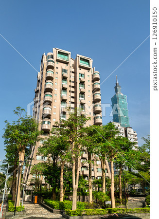 A bright morning view in Taipei, Taiwan, shows a tan residential building with rounded balconies rising beside a modern skyscraper, both emerging above a small park of slender trees and manicured 126910150