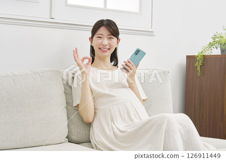Pregnant woman holding smartphone OK sign living room Pregnant woman holding smartphone OK sign living room 126911449