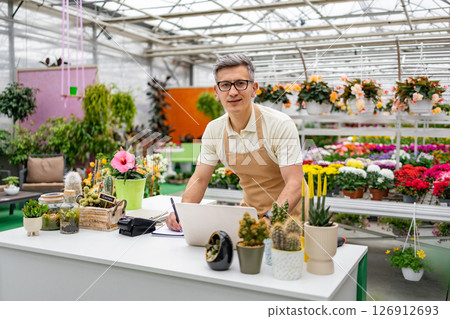 A Caucasian man works at a flower shop's counter, surrounded by a vibrant display of plants and flowers. 126912693