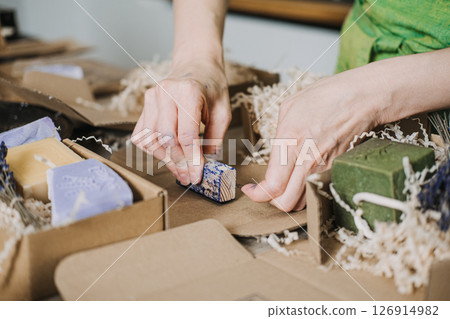 Person stamps a handmade soap bar with a logo inside kraft packaging filled with shredded filler. Handmade soap, logo stamping, artisanal branding, small batch production Person stamps a handmade soap bar with a logo inside kraft packaging filled with shredded filler. Handmade soap, logo stamping, artisanal branding, small batch production 126914982