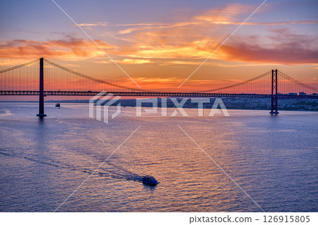 Iconic 25 de Abril bridge in silhouette over Tagus River, Lisbon, Portugal, at sunset with small boat sailing on calm waters. Vibrant orange and purple sky, dramatic backdrop, famous Portuguese 126915805