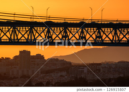 Striking close-up of famous 25 de Abril bridge, its silhouette against bright orange sunset sky. Lisbon, Portugal, cityscape and distant hills in hazy summer evening, blurred vehicles cross bridge 126915807