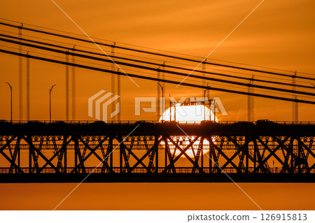 Glowing sun setting behind 25 de Abril bridge, Lisbon, Portugal, suspension cables, cars and bridge structure in silhouette. Bright orange sky, dramatic urban sunset over Tagus River. Close view Glowing sun setting behind 25 de Abril bridge, Lisbon, Portugal, suspension cables, cars and bridge structure in silhouette. Bright orange sky, dramatic urban sunset over Tagus River. Close view 126915813