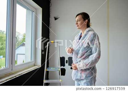 Woman in protective coveralls standing in front of unpainted wall with paint samples. Person preparing to paint interior wall at home. Concept of home renovation and interior upgrade Woman in protective coveralls standing in front of unpainted wall with paint samples. Person preparing to paint interior wall at home. Concept of home renovation and interior upgrade 126917339