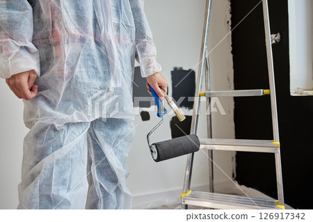 Woman wearing protective coveralls holds black paint roller near painted wall samples and ladder in room under renovation. Painter preparing to paint wall. Home improvement and interior update concept 126917342