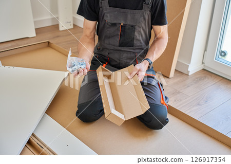 Man in work overalls opens plastic bag with screws and fittings while kneeling beside boxed furniture panels and tools. Furniture assembly Man in work overalls opens plastic bag with screws and fittings while kneeling beside boxed furniture panels and tools. Furniture assembly 126917354