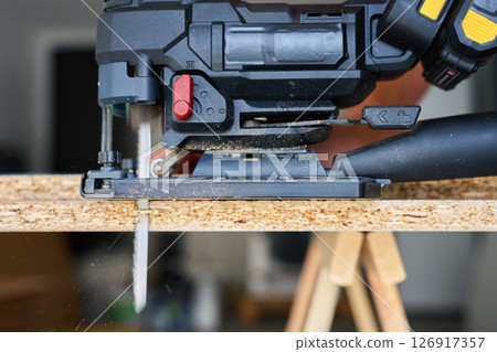Close up of electrical handheld jigsaw cutting wooden board in workshop. Work tools for manual labor. Carpentry and woodworking concept Close up of electrical handheld jigsaw cutting wooden board in workshop. Work tools for manual labor. Carpentry and woodworking concept 126917357