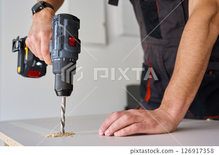 Male hands drilling wooden board with power drill. Process of carpentry working in home workshop, drills hole in chipboard Male hands drilling wooden board with power drill. Process of carpentry working in home workshop, drills hole in chipboard 126917358