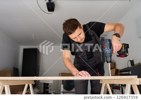 Man in work overalls bib using cordless power drill to make hole in laminated wood board. Male carpenter working in home workshop, drilling chipboard on workbench 126917359