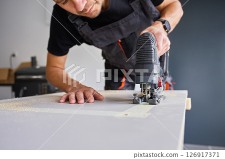 Worker in workwear using handheld cordless electric jigsaw to cut wooden board in garage. Skilled carpenter working in workshop. Man doing DIY home renovation 126917371
