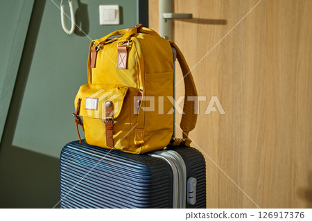 Packed dark suitcase and yellow backpack stand by door. Luggage ready for departure. Preparation for trip or travel Packed dark suitcase and yellow backpack stand by door. Luggage ready for departure. Preparation for trip or travel 126917376