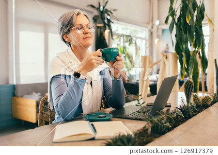 Senior woman with gray hair enjoying peaceful coffee break in cafe with laptop and notebook on table 126917379