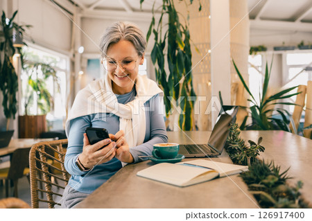 Senior woman with gray hair using smartphone in cafe, with laptop, coffee and notebook on the table Senior woman with gray hair using smartphone in cafe, with laptop, coffee and notebook on the table 126917400