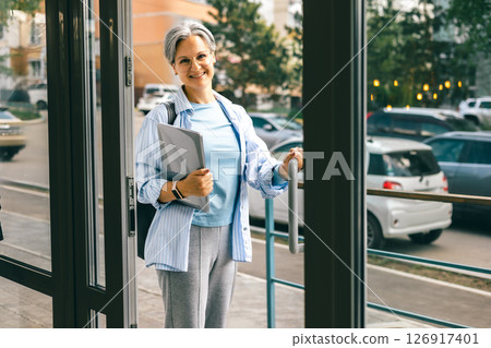 Stylish senior woman with laptop opening glass door and smiling on city street background in summer Stylish senior woman with laptop opening glass door and smiling on city street background in summer 126917401