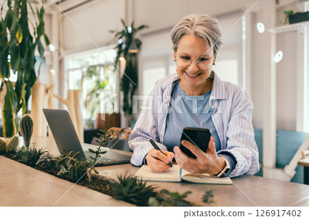 Smiling senior woman with gray hair writing notes while using smartphone at cozy modern workspace Smiling senior woman with gray hair writing notes while using smartphone at cozy modern workspace 126917402