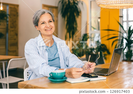 Smiling mature woman freelancer working on laptop and taking a break at cozy cafe workspace Smiling mature woman freelancer working on laptop and taking a break at cozy cafe workspace 126917403