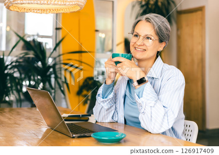 Midlife freelance woman smiling during coffee break while working from modern cozy workspace Midlife freelance woman smiling during coffee break while working from modern cozy workspace 126917409