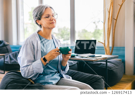 Peaceful senior woman with gray hair enjoying coffee break and mindfulness in cozy workspace 126917413