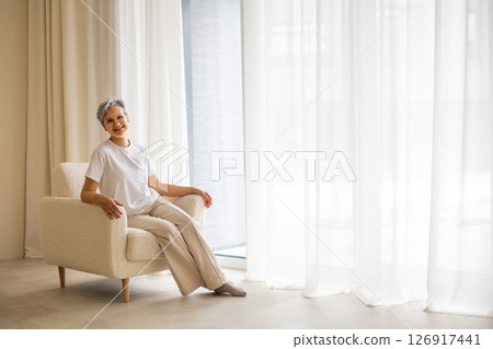 A mature elderly, well-groomed, beautiful woman pensioner with gray hair sits in a stylish chair 126917441