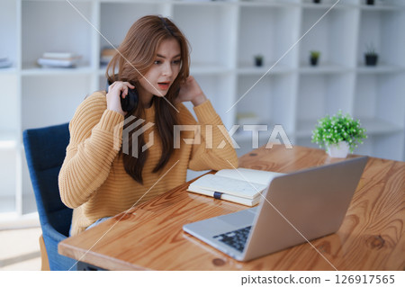 A young woman puts on headphones while working from home on her laptop. 126917565