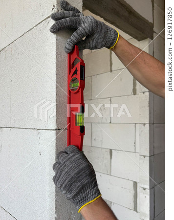 A close-up photo of a level in the hands of a builder measuring the evenness of a wall. A side view of the level on the wall. A close-up photo of a level in the hands of a builder measuring the evenness of a wall. A side view of the level on the wall. 126917850
