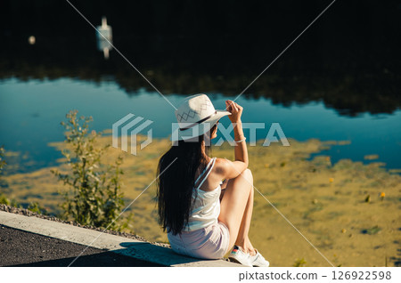A girl in white shorts and a T-shirt sits by the lake, immersed in the tranquility of nature 126922598