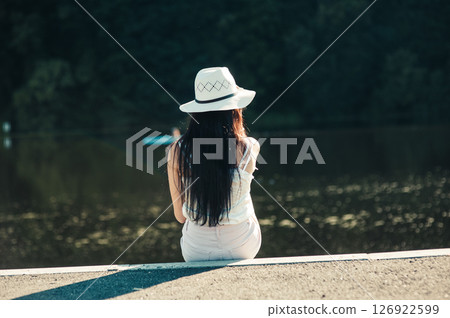 A girl in white shorts and a T-shirt sits by the lake, immersed in the tranquility of nature 126922599