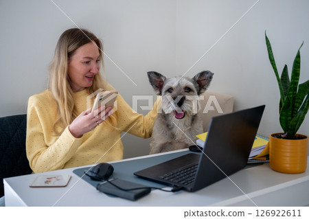 Woman in yellow sweater using phone with dog beside her at home office desk, concept of remote work Woman in yellow sweater using phone with dog beside her at home office desk, concept of remote work 126922611