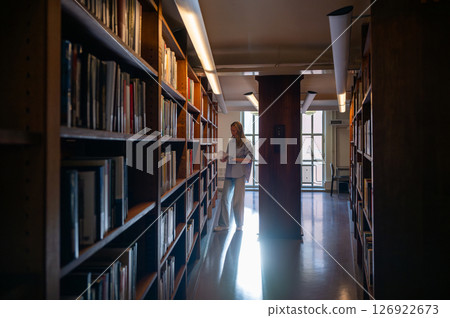 Woman reading a book in quiet library aisle with warm natural light Woman reading a book in quiet library aisle with warm natural light 126922673