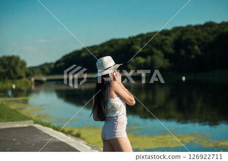 A slender girl by the water, dressed in a T-shirt and shorts, holds a phone and talks A slender girl by the water, dressed in a T-shirt and shorts, holds a phone and talks 126922711