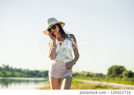 A girl in a white hat is standing by the pond and smiling sincerely while holding the phone A girl in a white hat is standing by the pond and smiling sincerely while holding the phone 126922716