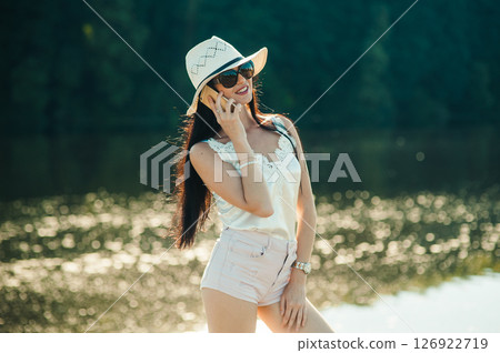 A girl in a white hat is standing by the pond and smiling sincerely while holding the phone A girl in a white hat is standing by the pond and smiling sincerely while holding the phone 126922719