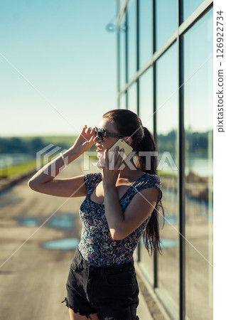 A young woman in a T-shirt and shorts is talking on the phone, standing next to a mirrored facade A young woman in a T-shirt and shorts is talking on the phone, standing next to a mirrored facade 126922734