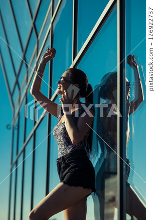 A young woman in glasses and a T-shirt is talking on the phone next to the transparent facade A young woman in glasses and a T-shirt is talking on the phone next to the transparent facade 126922737