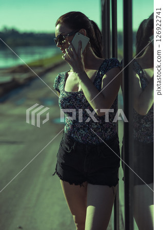 A girl in a stylish outfit is standing near a transparent glass facade, talking on the phone A girl in a stylish outfit is standing near a transparent glass facade, talking on the phone 126922741
