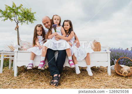 A young man embraces his charming daughters sitting next to him on a wooden retro bench in the lavender field 126925036