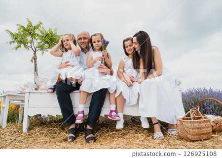 A happy family sits on a white wooden retro bench in the middle of a charming blooming field 126925108