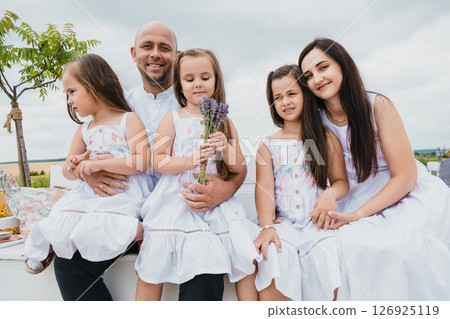 On a white wooden bench in the field, a father and mother sit with their three wonderful daughters 126925119