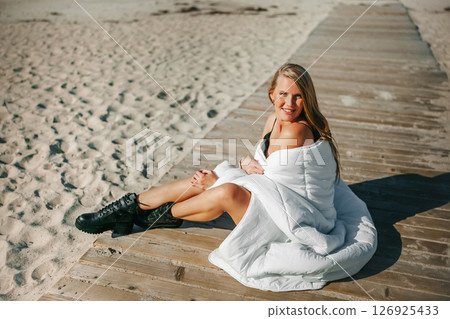 A serene beach moment with a woman wrapped in white blanket enjoying the sun on a wooden dock by the 126925433