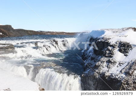 horizontal photo took in Gullfoss Falls, Hvita river, Golden Circle, Reykjavik, Iceland. A waterfall cascading over rocky cliffs into a river below with snow covered areas and a clear blue sky 126926979
