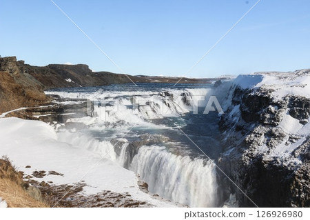 horizontal photo took in Gullfoss Falls, Hvita river, Golden Circle, Reykjavik, Iceland. A waterfall cascading over rocky cliffs into a river below with snow covered areas and a clear blue sky 126926980