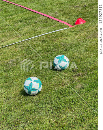Soccer balls placed on a grassy field next to a training cone setup, ready for practice. The red cone and white line marker indicate a training zone, enhancing sports training sessions. Soccer balls placed on a grassy field next to a training cone setup, ready for practice. The red cone and white line marker indicate a training zone, enhancing sports training sessions. 126927811