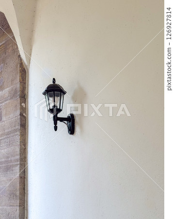 Classic black wall mounted lantern provides elegant lighting against a stone wall. The traditional fixture enhances the timeless architecture, casting soft shadows on the surface. Classic black wall mounted lantern provides elegant lighting against a stone wall. The traditional fixture enhances the timeless architecture, casting soft shadows on the surface. 126927814